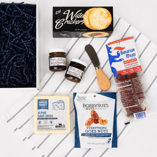 A curated gourmet snack and cheese gift set arranged on a white tray with thin gray stripes. Visible items include a box of Elk Water Crackers, two small artisanal spreads in jars, a small knife, a package of Bavarian Meals hot dogs, a package of Bobbysue's Everything Goes Nuts, and a block of Farmer's Market Alpine Baby Swiss cheese.