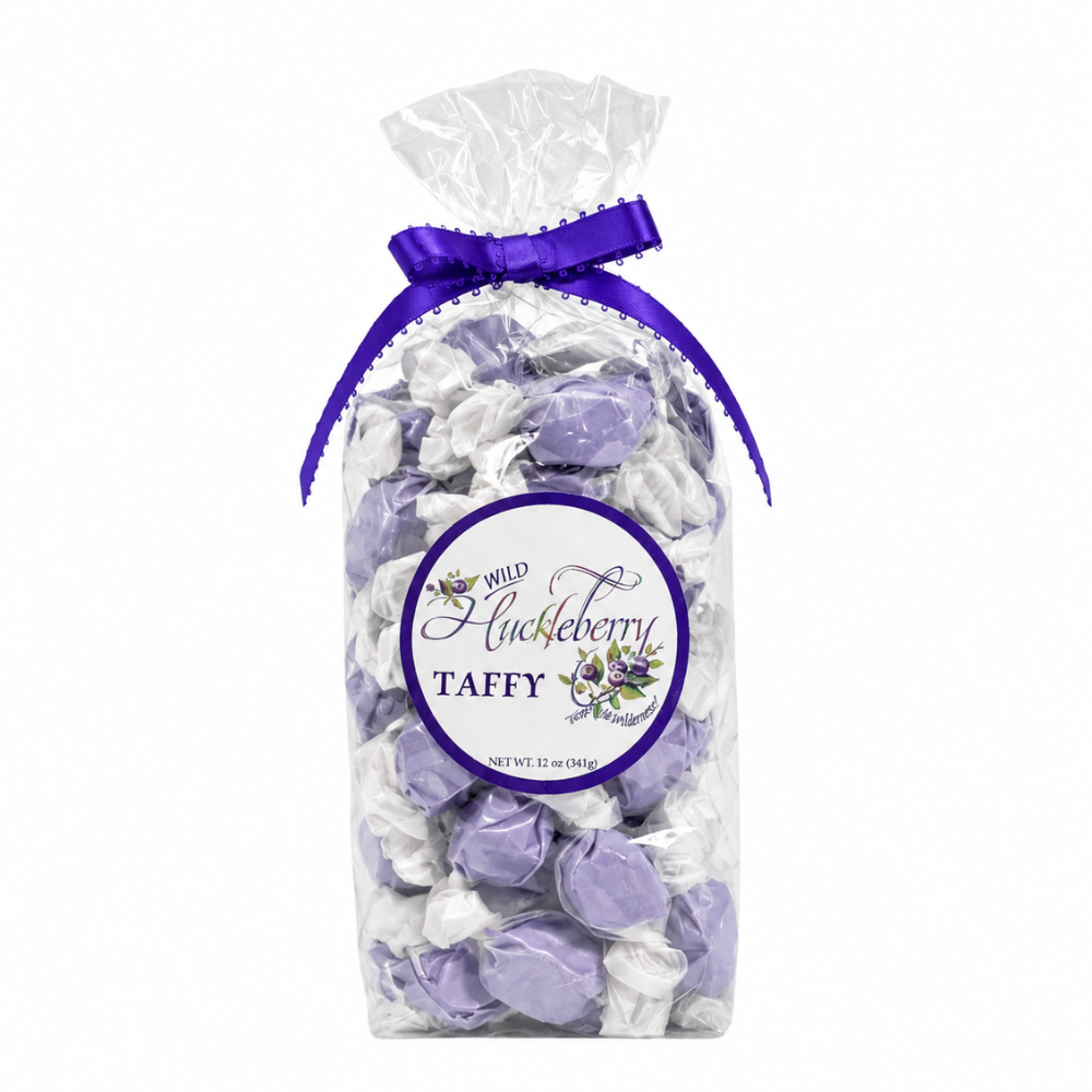 A clear cellophane bag filled with wrapped taffy candies in light purple and white, tied with a purple ribbon at the top. A round front label reads 'Wild Blueberry Taffy' with floral graphics.