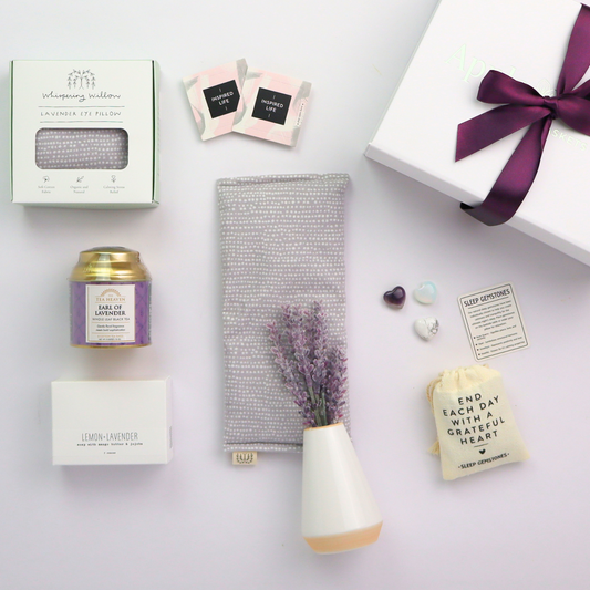 Collection of wellness products including a candle, eye pillow, and lavender sachet on a light surface. Unboxed Lavender Self-Care Gift Box items including tea tin, soap, sleep stones, pop-open cards, eye pillow, and bud vase