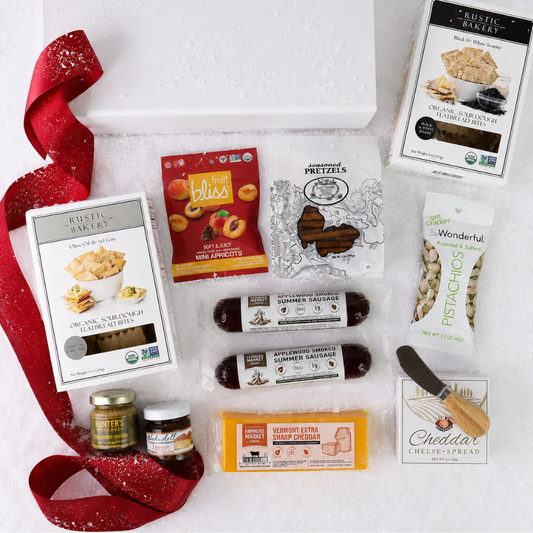 A flat-lay gift basket of gourmet snacks and cheeses on a white background with a white basket, a red glitter ribbon, and multiple packaged food items including Rustic Bakery items, Bliss candy, pretzels, pistachios, sausage, cheddar cheese, and a small knife.