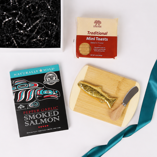 A styled flat-lay gift set on a white background featuring a box of smoked salmon, Divine traditional mini toasts, a wooden cutting board with a small knife, and a teal ribbon.