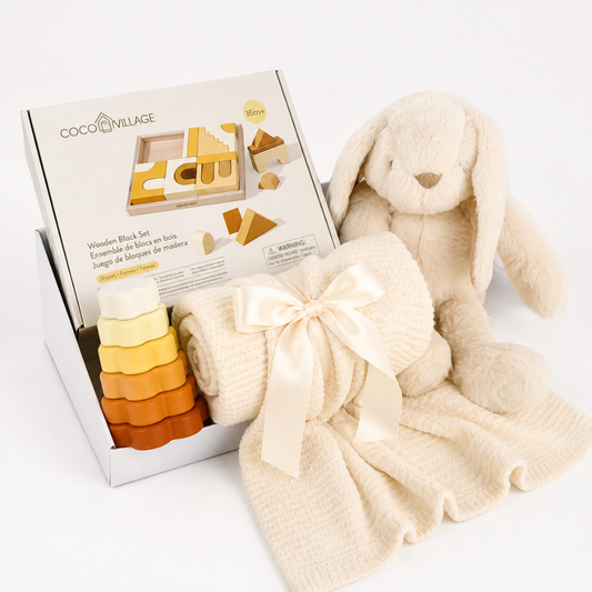 Coco Village wooden blocks set in a white display box, styled with a cream bunny plush toy, a cream waffle-knit blanket tied with a satin bow, and a small stack of wooden blocks in cream, yellow, and orange on a white background.