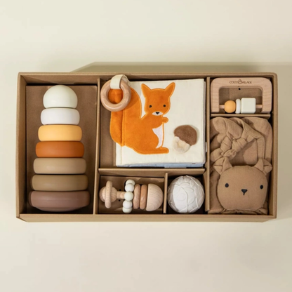 Neutral baby gift box with wooden rattle, silicone stacking tower, lovey, and woodland sensory book arranged in a kraft box.