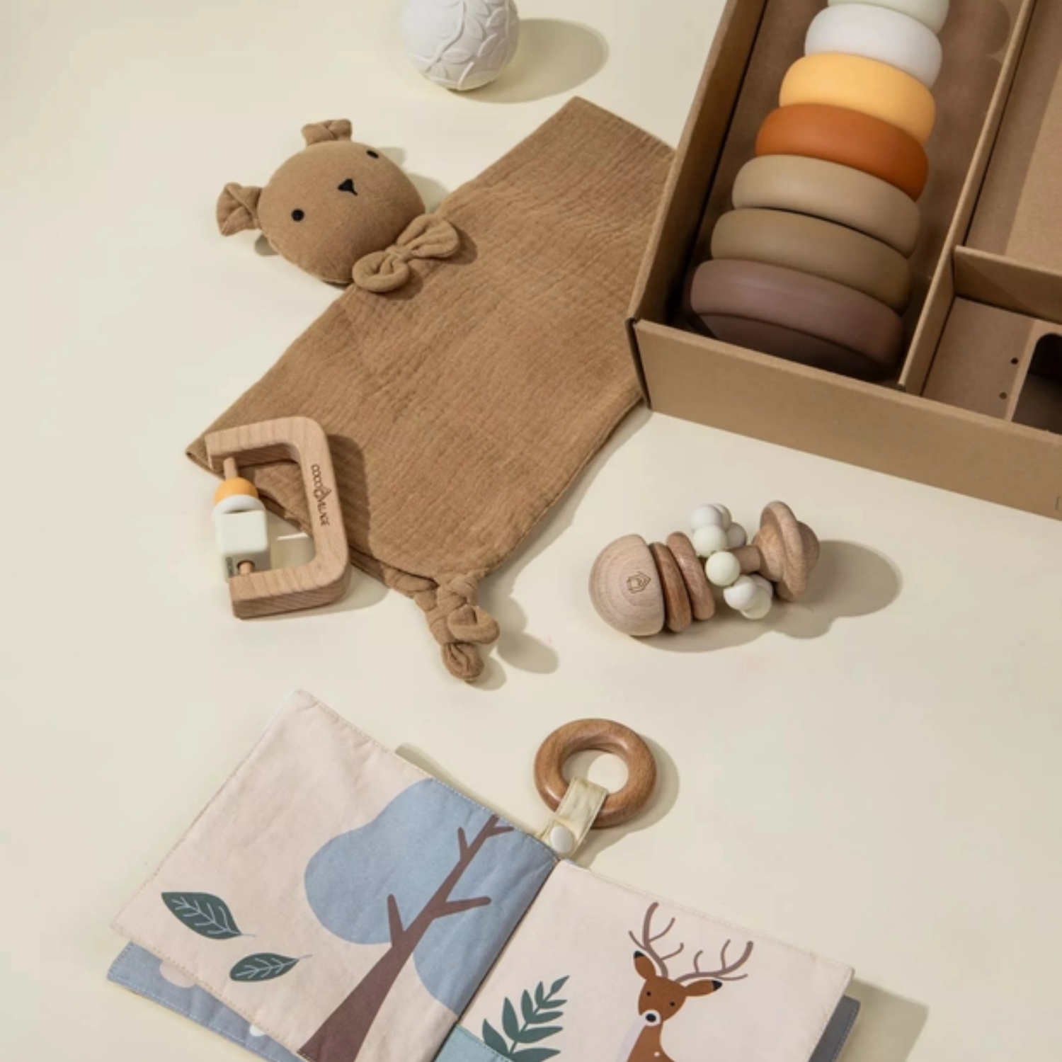 Flat lay of modern baby toys including a lovey, silicone stacking tower, wooden rattle, teether, and woodland sensory book in warm neutral tones.