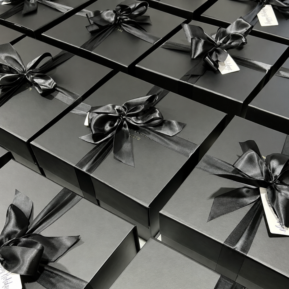 Multiple black gift boxes arranged in a grid, each tied with a black satin ribbon and bow and featuring a small white tag.