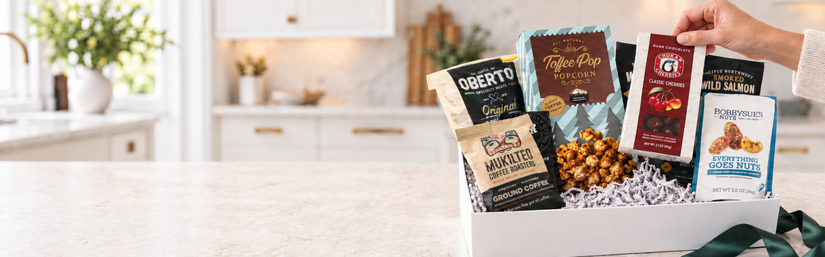 A curated gourmet gift box filled with assorted washington snack and coffee packages, held by a person's hand in a bright, modern kitchen setting.