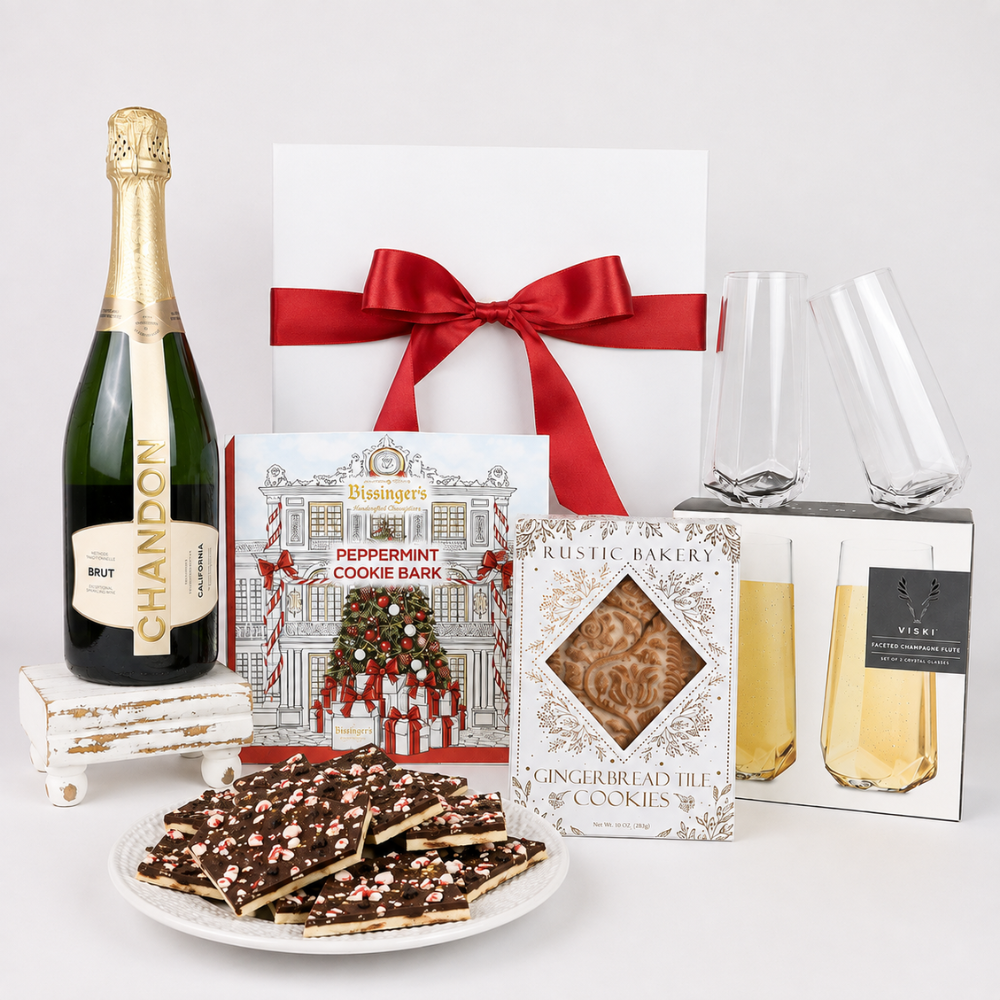 A luxury gift hamper set featuring a bottle of Chandon Brut, a white gift box with a red ribbon, a plate of peppermint chocolate cookie bars, a box of Rustic Bakery gingerbread tin cookies, a peppermint cookie bark box, two champagne flutes, and a boxed Viski brand drink, all arranged on a white background.