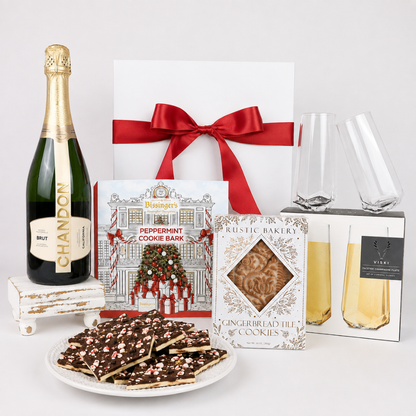 A luxury gift hamper set featuring a bottle of Chandon Brut, a white gift box with a red ribbon, a plate of peppermint chocolate cookie bars, a box of Rustic Bakery gingerbread tin cookies, a peppermint cookie bark box, two champagne flutes, and a boxed Viski brand drink, all arranged on a white background.