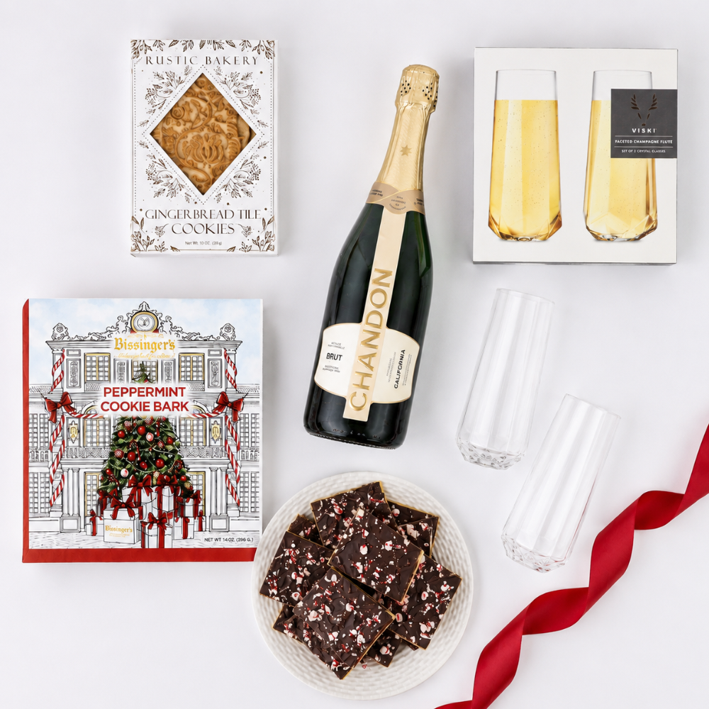 A curated gift set arranged on a white background, including a bottle of Chandon sparkling wine, a box of Rustic Bakery gingerbread cookies, a Biesinger's peppermint cookie bark box, a plate of chocolate cookie bites with sprinkles, two champagne flutes in packaging, and a red ribbon.