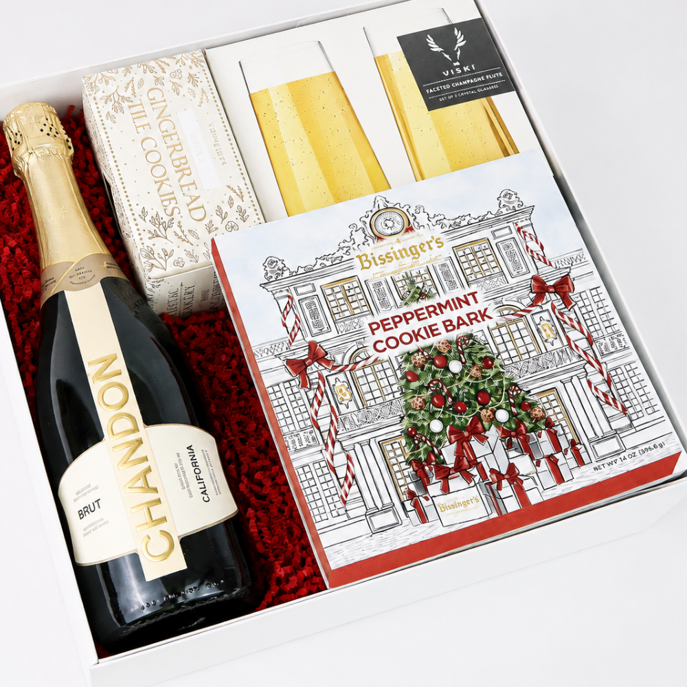 A gift box arrangement featuring a bottle of Chandon Brut, a box of Bisinger's Peppermint Cookie Bark, two gold champagne flutes, and a gingerbread tile cookie, all arranged in red foam inside a white gift box.