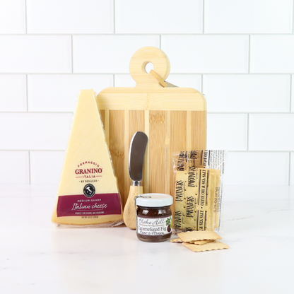 Bamboo cutting board with cheese, knife, and other items on a white surface.