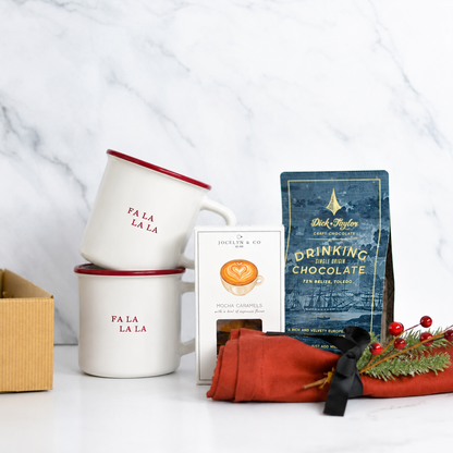 A holiday gift set arranged on a white marble surface, including two stacked white mugs with red rims and red 'FA LA LA' text, a box of Dr. Johnson & Co.
