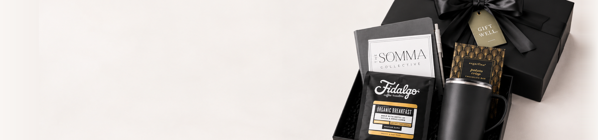 A premium gift set featuring a black box with a black ribbon, a black card from ISOMMA Lifestyle, a bottle of Tidalgo body wash, and a black cylindrical container, arranged on a clean white background with ample negative space.