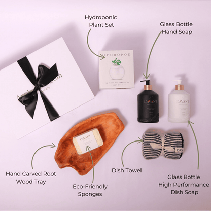 Flat lay of high-end home gift set, showcasing L'AVANT Collective home care products, an acacia wood board, hydroponic herb kit, soft kitchen towels, and sustainable sponges—ideal for housewarming gifting.