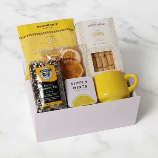 A Drop of Joy lemon-themed snack box filled with lemon shortbread cookies, a yellow ceramic teacup, citrus crisps, loose tea, lemon drops, and mints—perfect for a housewarming gift basket or self-care gift box.
