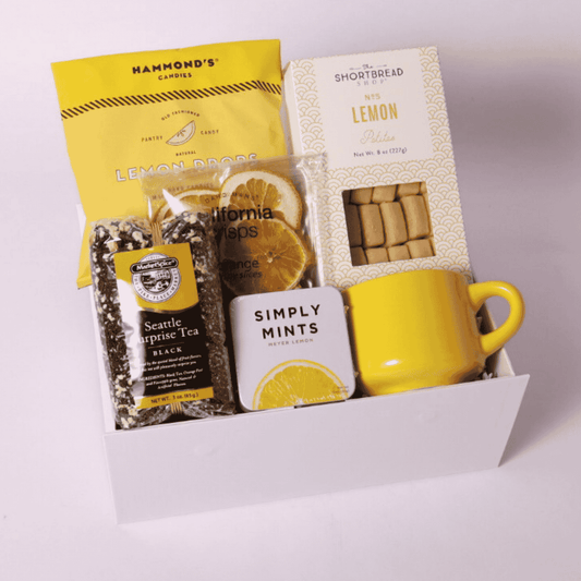 A Drop of Joy lemon-themed snack box filled with lemon shortbread cookies, a yellow ceramic teacup, citrus crisps, loose tea, lemon drops, and mints—perfect for a housewarming gift basket or self-care gift box.