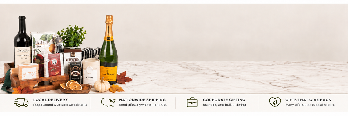 A wide promotional banner for a pacific northwest gift basket service. The left side shows a curated gift arrangement on a marble surface with a wine bottle, champagne bottle, small plant, seasonal decorations, and gift boxes.