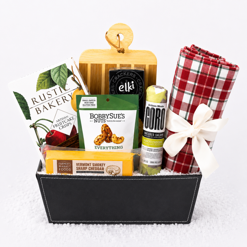 A curated gift basket in a black rectangular tray filled with assorted food items and a rolled red-and-green plaid blanket tied with a white bow. Visible products include a bag of Rustic Bakery cinnamon fruitcake crisp, an Elk Creek crackers pack, a package of Bobby Sue's Everything nuts, a bar of chocolate, a tube of CORO mint tea, and a wooden cutting board.