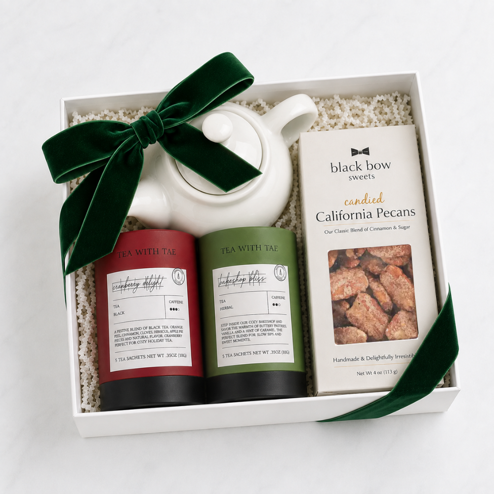 A white gift box filled with tea and sweets, tied with a dark green velvet bow. The box contains a white teapot, two tea tins labeled 'Tea With Tai' in red and green, and a box of 'Black Bow Sweets' featuring 'Candied California Pecans'.