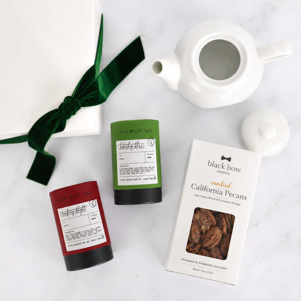 A styled flat-lay of a gift set on a white marble surface, featuring a white teapot, a white gift box with a dark green velvet bow, a red cylindrical tea canister, a green cylindrical tea canister, and a white box of Black Bow Foods California Pecans.