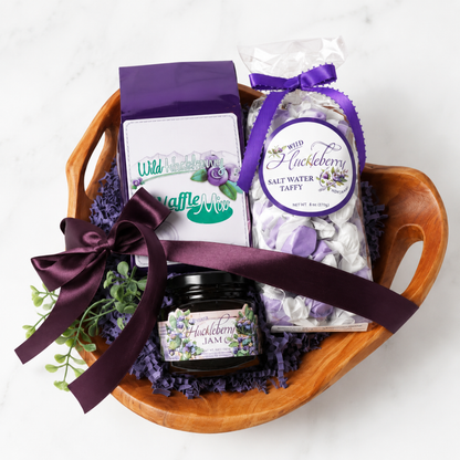 A huckleberry -themed gift basket arranged in a wooden bowl, featuring a bag of Wild Huckberry Waffle Mix, a bag of Wild Huckberry Salt Water Taffy, a jar of Wild Huckberry Jam, and a purple ribbon, set on a white marble background.