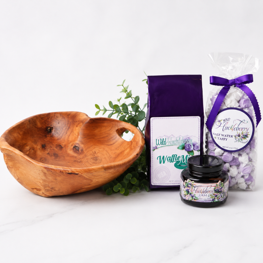 A gift set featuring a carved wooden bowl, a purple bag of waffle mix, a jar of huckleberry waffle mix, and a bag of huckleberry salt water taffy on a white marble surface.