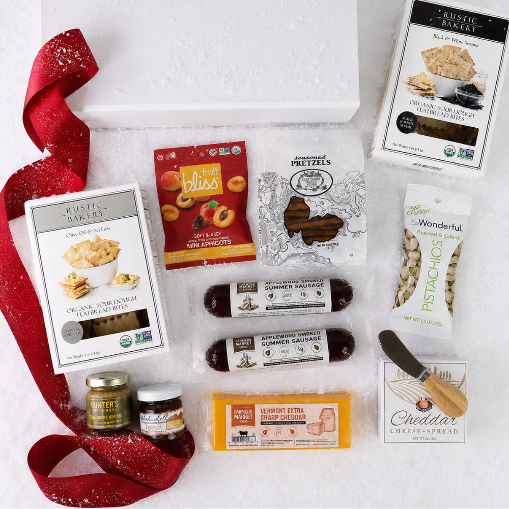 A flat-lay gift basket of gourmet snacks and cheeses on a white background with a white basket, a red glitter ribbon, and multiple packaged food items including Rustic Bakery items, Bliss candy, pretzels, pistachios, sausage, cheddar cheese, and a small knife.