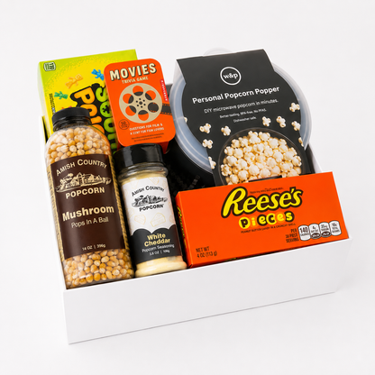 A curated popcorn gift set arranged in a white box, featuring a jar of Amish Country Popcorn Mushroom Pops in A Ball, a jar of Amish Country Popcorn White Cheddar Pops, a black wrap Personal Microwave Popcorn Popper, a box of Reese's Pieces, and a green and orange 'Movies for the Home' box.
