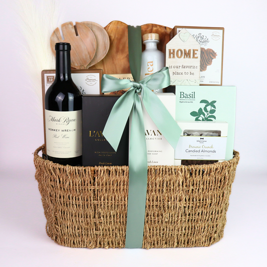 Luxury housewarming gift basket with wine, artisan hand soap, lotion, cutting boards, crackers, almonds, and a basil kit, displayed in a woven basket with a sage green satin bow.