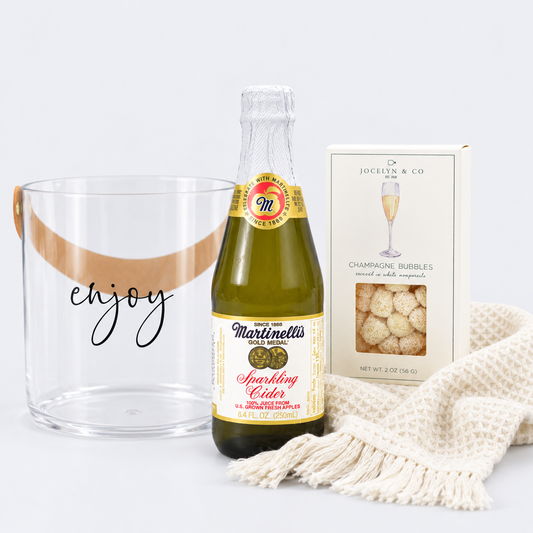 A sparkling cider gift set featuring a bottle of Martinelli Sparkling Cider, a clear glass bucket with a tan handle and the word 'enjoy' in black script, and a box of Jocelyn & Co Champagne Bubbles, styled with a cream knit throw blanket on a white background.