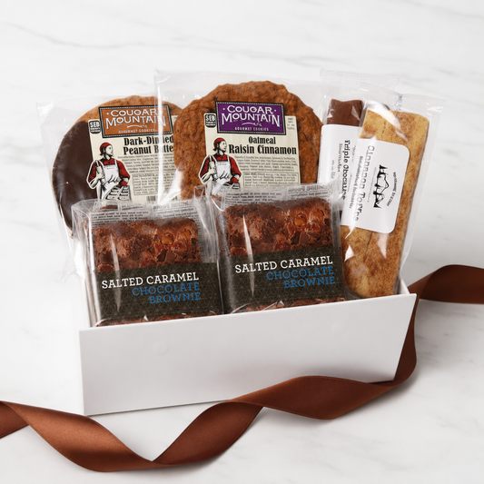 A gift box of cookies and brownies from Cougar Mountain Bakes, including Dark-Chocolate Peanut Butter Cookies, Oatmeal Raisin Cinnamon Cookies, Salted Caramel Chocolate Brownies, and a Chocolate Chip Cookie, arranged in a white box with a brown ribbon on a white marble background.