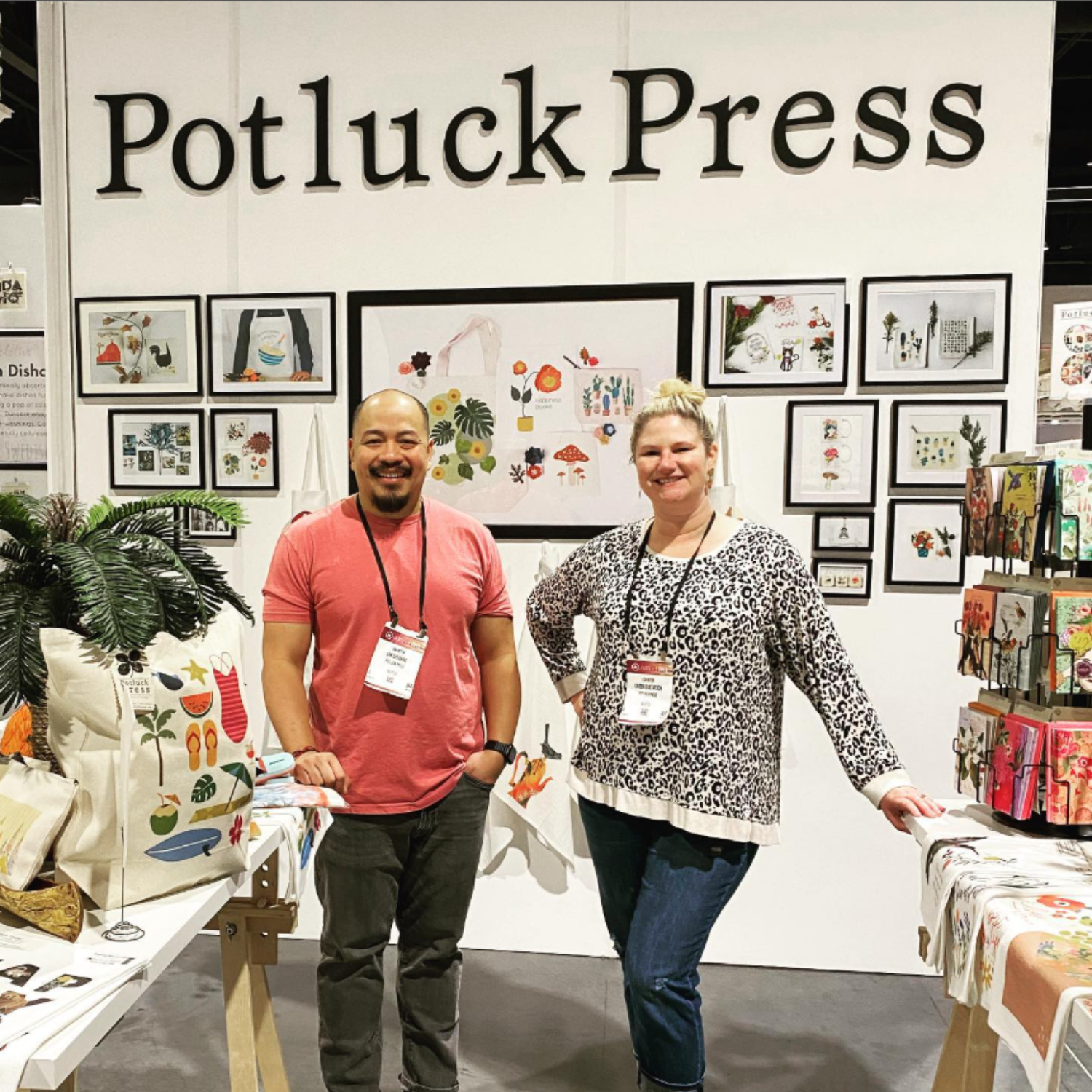 Two people standing in front of a 'Potluck Press' booth with various products on display.