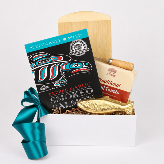 A gift box set featuring a package of Naturally Wild Pepper Garlic Smoked Salmon, a box of Bunk Traditional Mini Toasts, a wooden paddle, and a gold fish-shaped ornament in a white box with a teal ribbon, set against a plain white background.