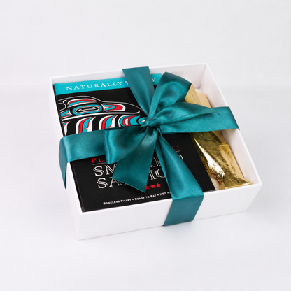 A white gift box tied with a large teal satin bow, containing a black packaged food item with colorful graphic artwork and a gold foil-wrapped item, photographed on a clean white background.