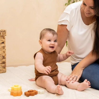 Baby in a brown onesie sitting on a bed with a woman, surrounded by toys.