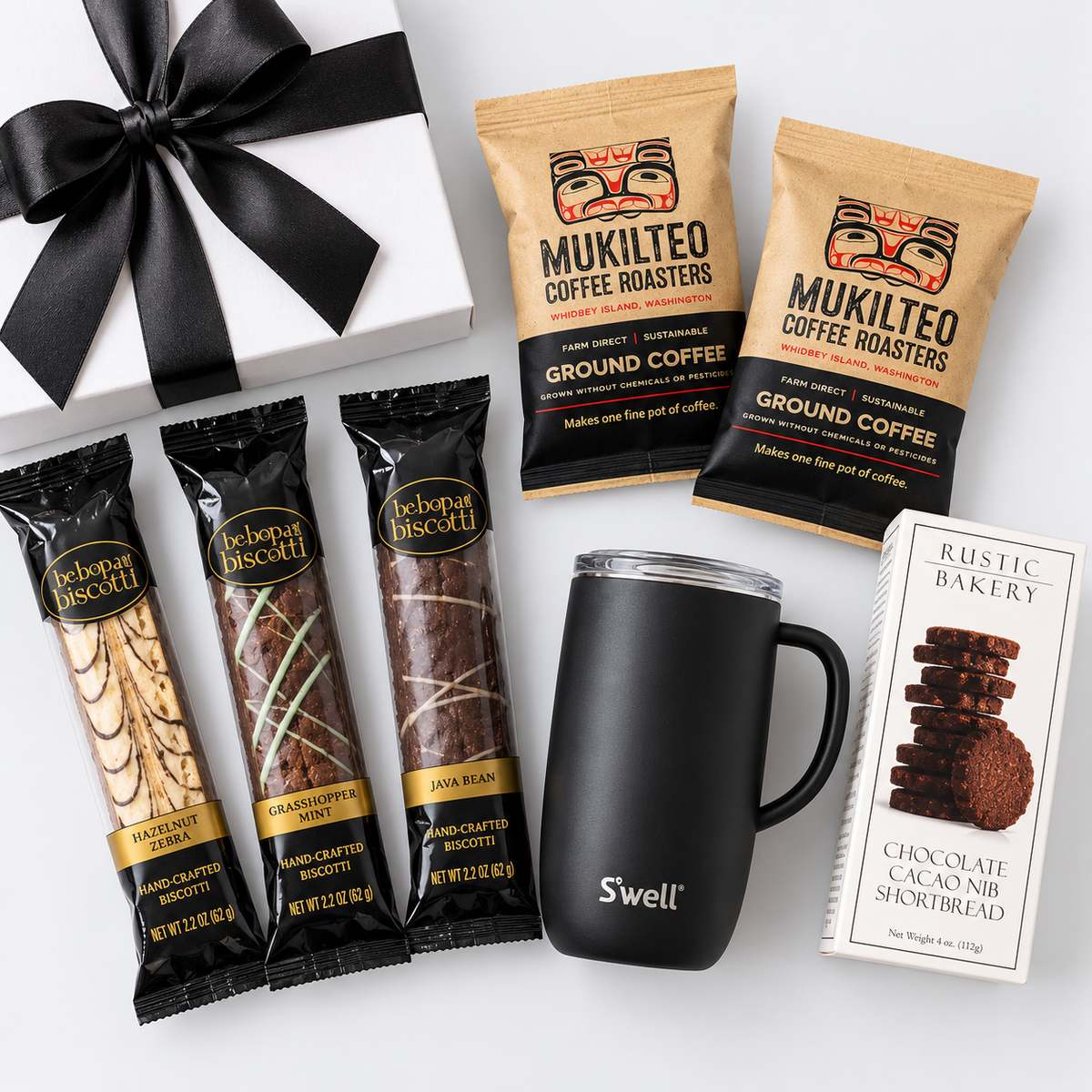 A gift set featuring a white gift box with a black ribbon, three Mukilteo coffee roaster bags, three Mukilteo biscuit bars, a black Swell travel mug, and a box of Aesthetic Bakery chocolate cak nibs, arranged on a white background.