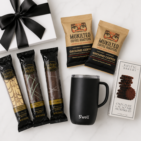 A gift set featuring a white gift box with a black ribbon, three Mukilteo coffee roaster bags, three Mukilteo biscuit bars, a black Swell travel mug, and a box of Aesthetic Bakery chocolate cak nibs, arranged on a white background.
