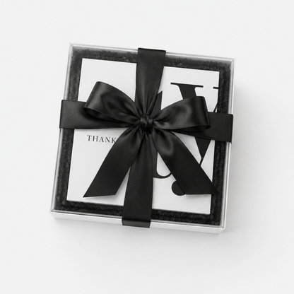 A square gift box with a clear lid, lined with black velvet, tied with a wide black satin ribbon bow. A white card with partial black lettering is visible inside.