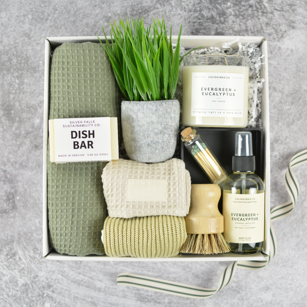 kitchen gift basket that includes a candle, dish towels, dish bar, plant, and bottles in a box on a gray background