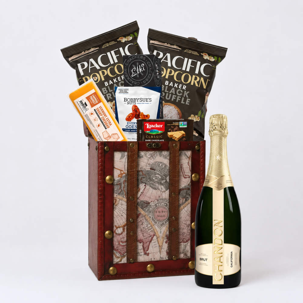 A gourmet gift basket set containing a bottle of Brut sparkling wine, bags of Pacific Popcorn and pacific popcorn, and a map-patterned wooden crate with brown leather straps.