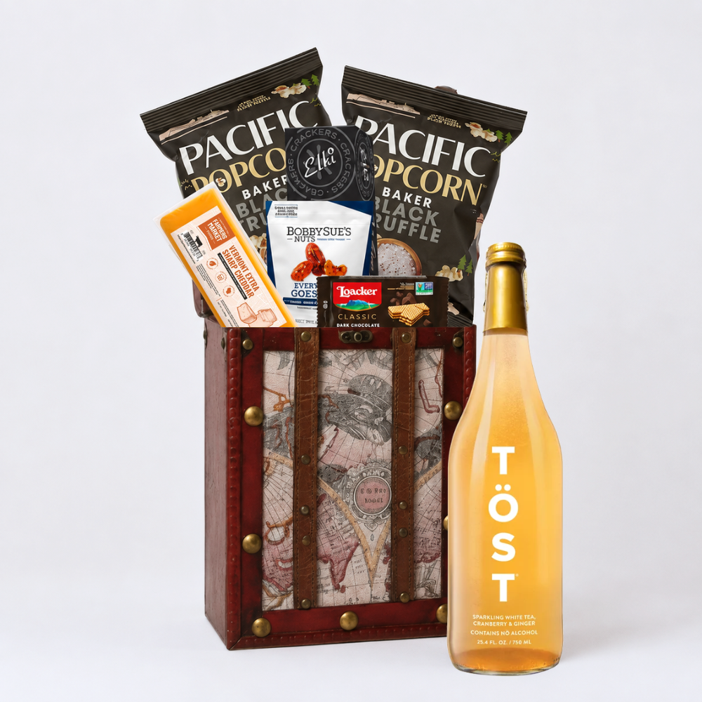 A curated gift basket or snack hamper set on a light gray studio background, featuring a wooden crate with vintage map artwork and leather straps, filled with snacks including bags of Pacific Popcorn, alongside a large bottle of Töst rosé wine.