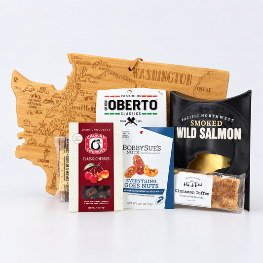 A curated gift basket or snack assortment arranged on a white studio background, including a wooden Washington state map board, a box of Oberto Classics, a package of Bobbys' Sues Nuts, a bag of smoked wild salmon, and a packaged cinnamon taffy.