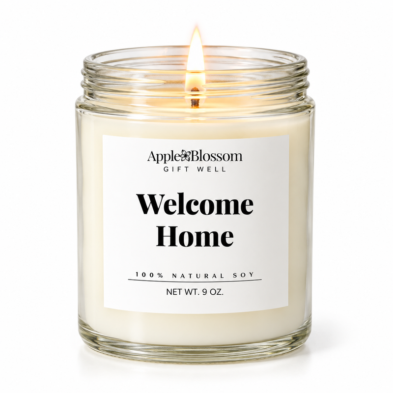 A lit 9 oz soy candle in a clear glass jar with a white label that reads 'Apple & Blossom Gift Well Welcome Home 100% Natural Soy'.