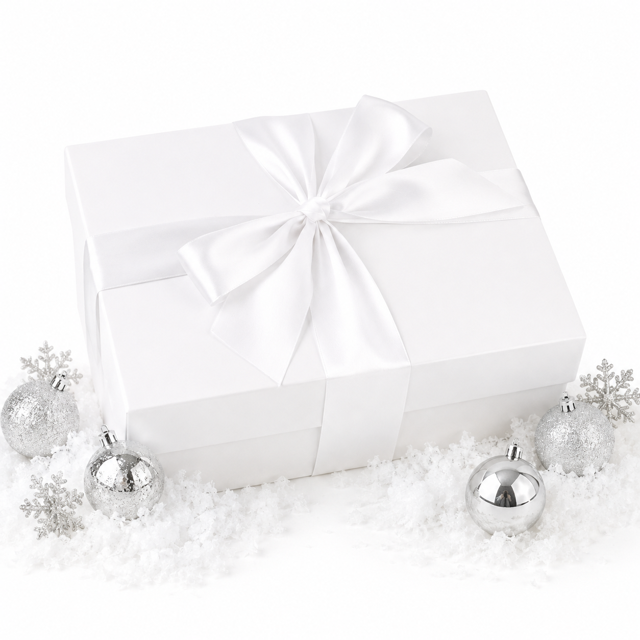A white gift box wrapped with a large white satin ribbon and bow, styled on a white surface with scattered faux snow and silver holiday ornaments.