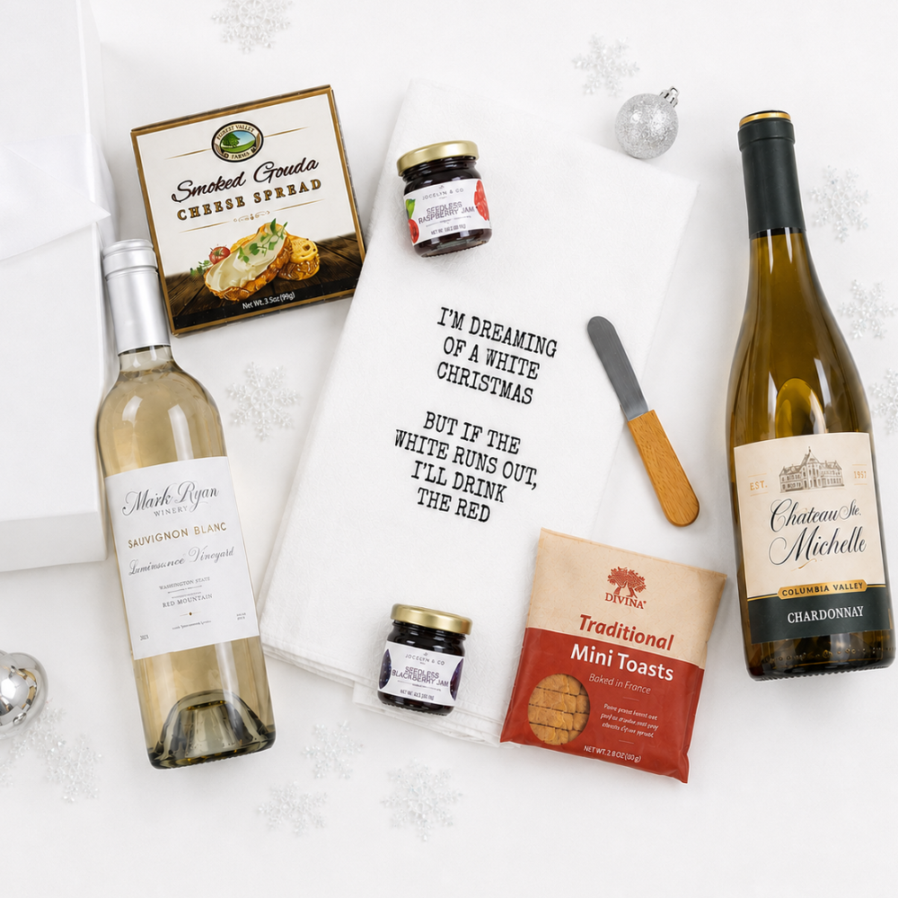A curated gift basket arrangement on a white background with snowflake graphics. The set includes a bottle of Musk Deer Sauvignon Blanc, a bottle of Chateau Michelle Chardonnay, a jar of Smoked Goula Cheese Spread, a jar of preserves, a bag of Chik-fil-A Traditional Mini Toasts, a white tea towel with black text, a butter knife, and a bag of mini breads.